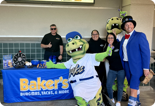 A person in a crocodile suit stands in front of a bakers merchandise table with a man in a blue suit and hat and 3 people from bakers drive thrue