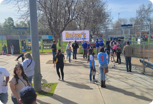 People standing in front of a sign for Bakers drive thru at a baseball game